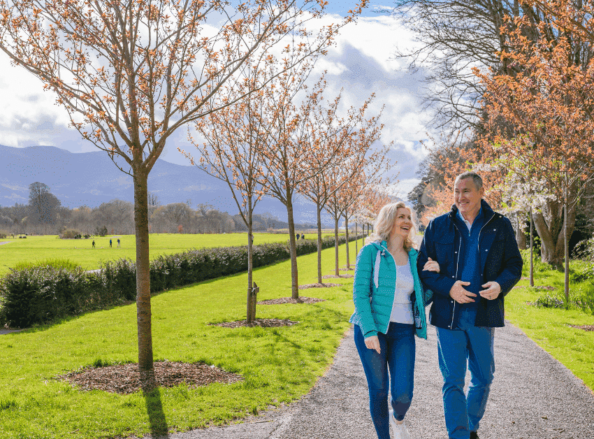 Couple strolling under blossoming trees along scenic mountain-view path next to a luxury hotel's garden.