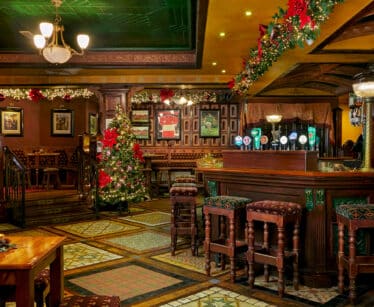 Cosy hotel pub with festive decor, ornate wooden bar, Christmas tree, comfortable seating, warm lighting.