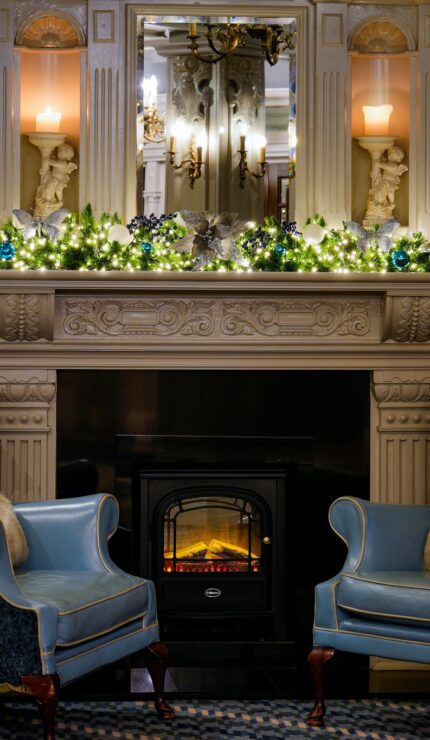 Cosy hotel lobby with elegant fireplace, festive lights, plush blue armchairs, inviting ambiance for relaxation and socialising.