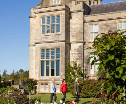 Guests stroll through vibrant gardens of a historic stone hotel with Gothic architecture under clear blue skies.