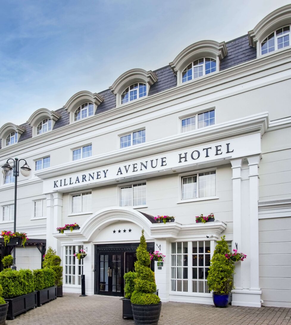 Elegant white facade of Killarney Avenue Hotel with lush greenery, inviting entrance, and charming vintage architecture.