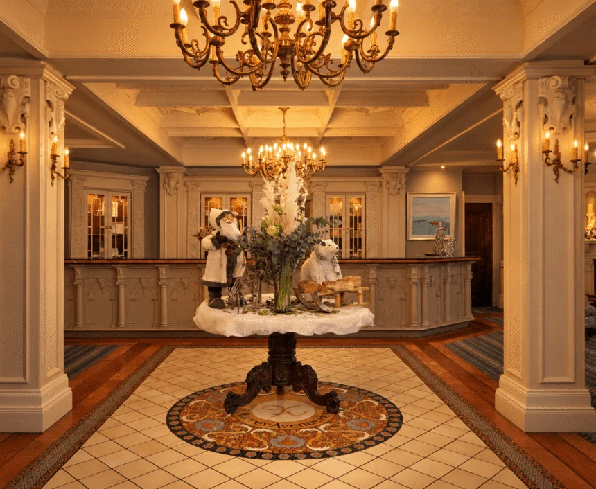 Elegant hotel reception with carved wooden desk, chandeliers, central floral display and festive decor, warm lounge seating visible